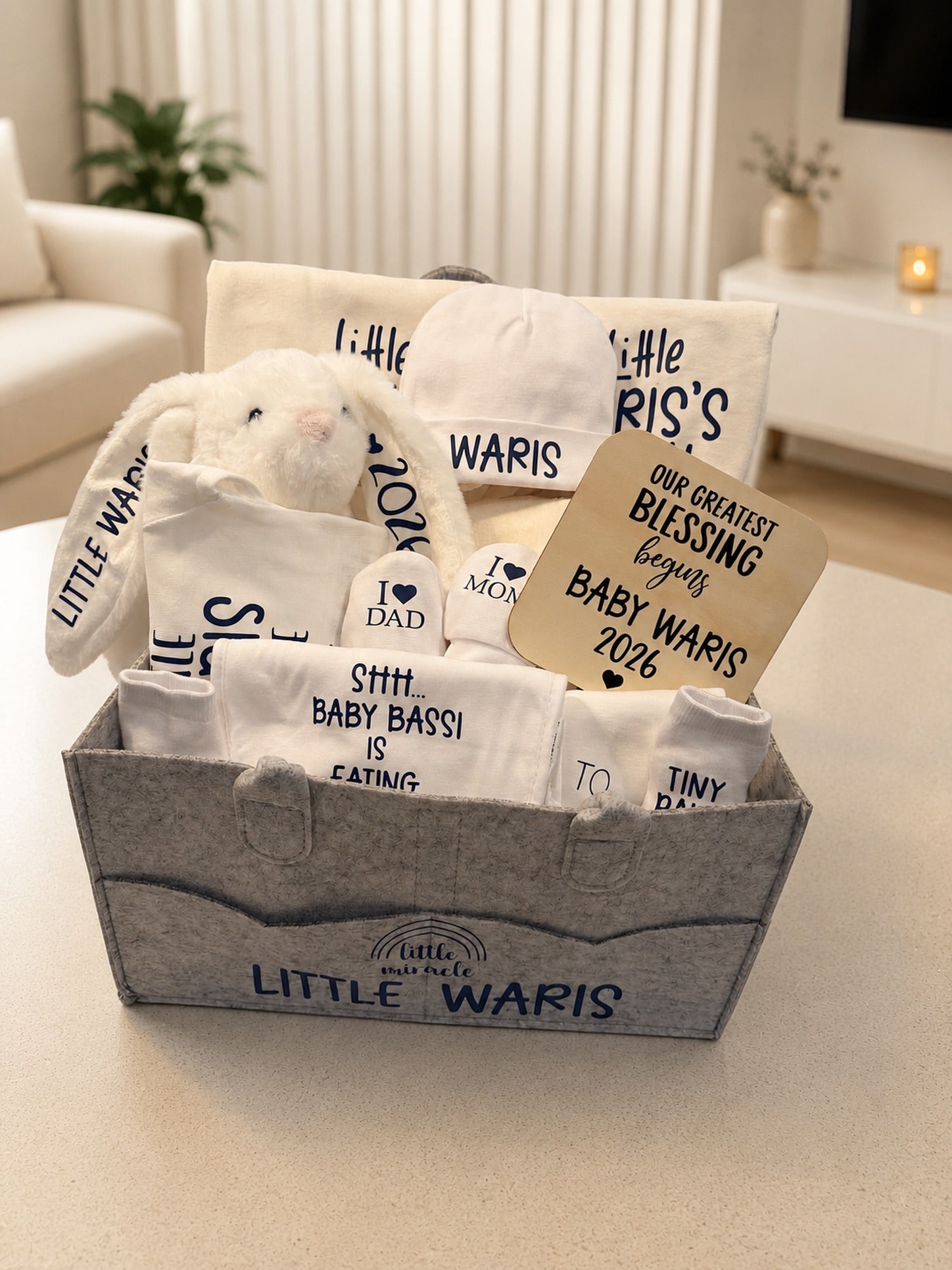 A curated baby gift basket set in a gray felt storage box, containing a plush white bunny, white baby clothes or blankets with black text prints, a kraft paper tag, and a small white bottle. The basket is labeled 'LITTLE WARIS' and sits on a light beige tabletop in a softly lit living room.